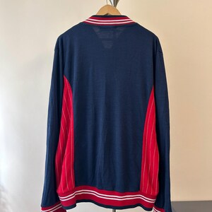 Puede incluir: Una chaqueta de manga larga azul marino con paneles laterales rojos y finas rayas verticales blancas. La prenda incluye cuello, pu&ntilde;os y dobladillo acanalados, cada uno detallado con dos rayas blancas horizontales sobre un fondo rojo.