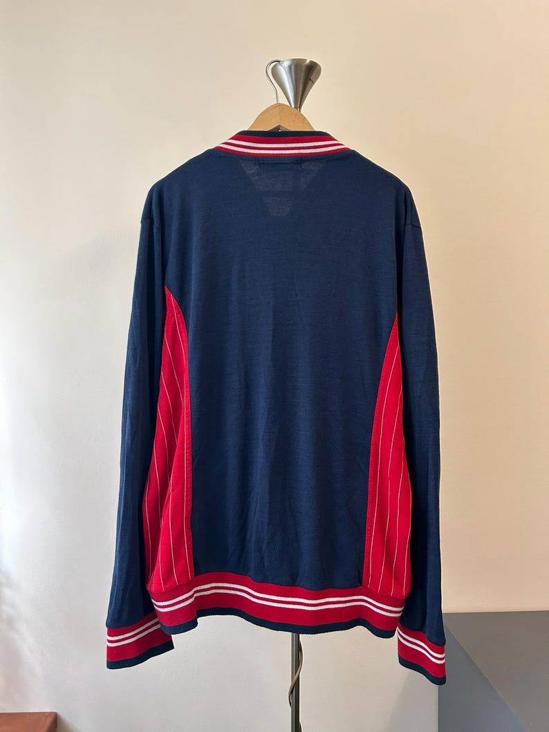 Puede incluir: Una chaqueta de manga larga azul marino con paneles laterales rojos y finas rayas verticales blancas. La prenda incluye cuello, pu&ntilde;os y dobladillo acanalados, cada uno detallado con dos rayas blancas horizontales sobre un fondo rojo.