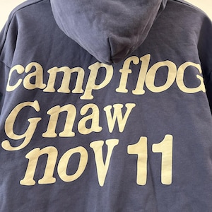 May include: A navy blue hooded sweatshirt with the words "campfloc Gnaw nov 11" printed in a cream-colored font. The hoodie has a drawstring and a kangaroo pocket.