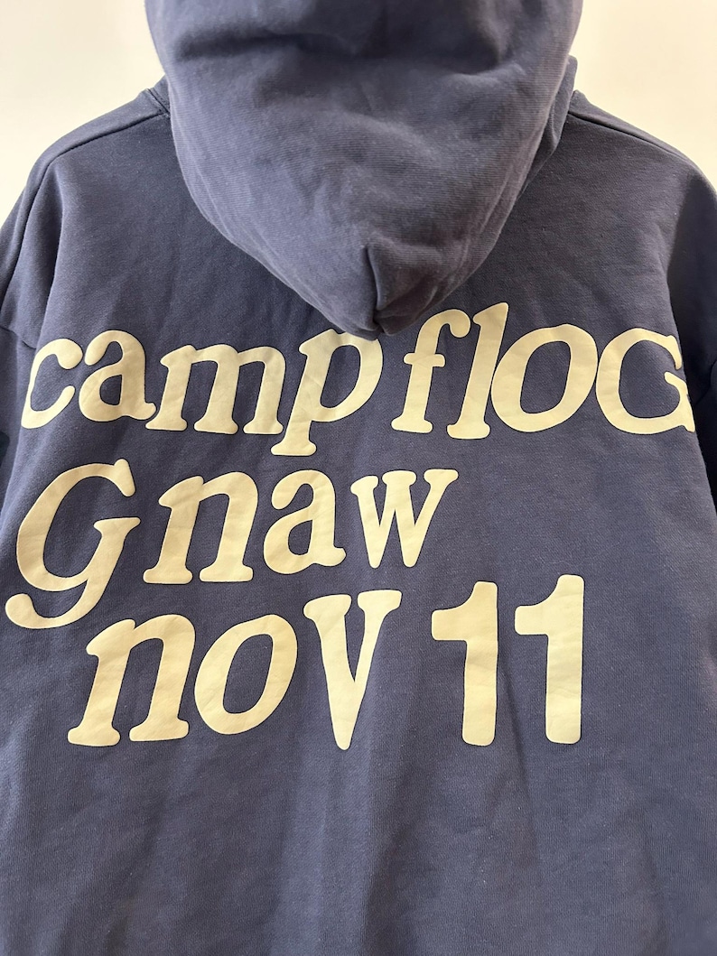 May include: A navy blue hooded sweatshirt with the words "campfloc Gnaw nov 11" printed in a cream-colored font. The hoodie has a drawstring and a kangaroo pocket.