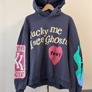 May include: Navy blue hoodie with the text "lucky me i see Ghosts" in cream-colored lettering. A pink heart with the word "feel" is centered on the chest. The left sleeve has a red and white patch with text, and the right sleeve has a flame design.