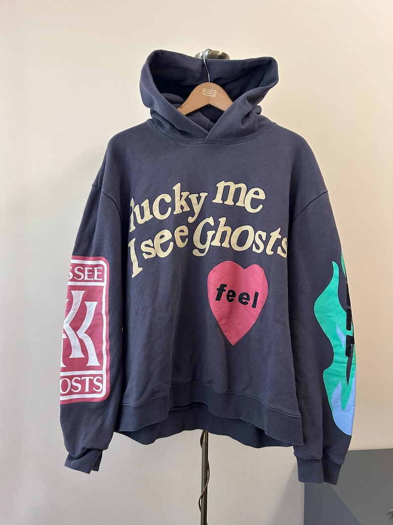 May include: Navy blue hoodie with the text "lucky me i see Ghosts" in cream-colored lettering. A pink heart with the word "feel" is centered on the chest. The left sleeve has a red and white patch with text, and the right sleeve has a flame design.