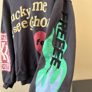 May include: Dark gray hoodie with the text "lucky me see ghosts" in yellow, a red heart with "fe" in white, and "FREE" in black with green and blue flames on the sleeve. The left sleeve has "SEE" and "GHOSTS" in red and white.