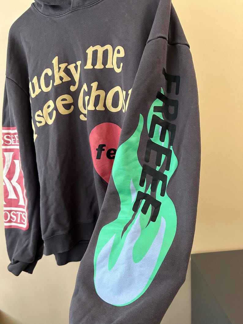 May include: Dark gray hoodie with the text "lucky me see ghosts" in yellow, a red heart with "fe" in white, and "FREE" in black with green and blue flames on the sleeve. The left sleeve has "SEE" and "GHOSTS" in red and white.