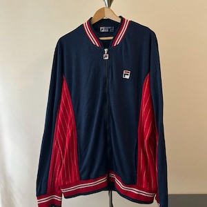 Puede incluir: Una chaqueta deportiva azul marino con cremallera y paneles laterales rojos con finas rayas blancas. La chaqueta tiene cuello, pu&ntilde;os y dobladillo acanalados con rayas rojas y blancas. Un logotipo de Fila est&aacute; bordado en el pecho izquierdo.