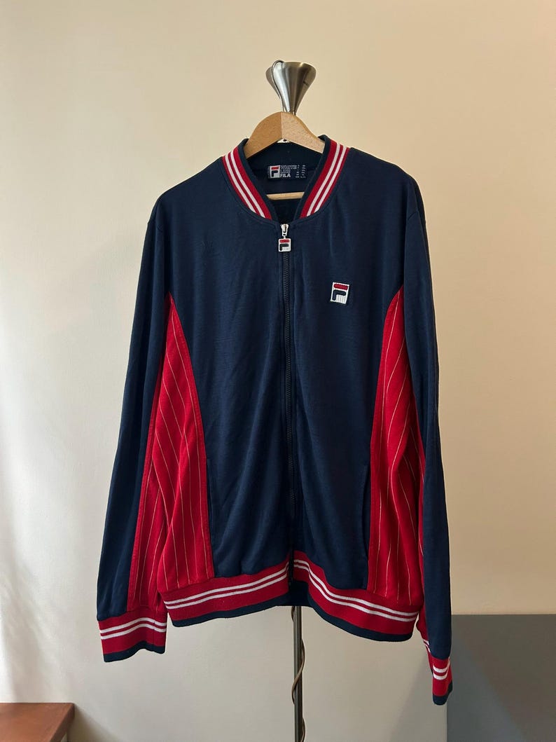 Puede incluir: Una chaqueta deportiva azul marino con cremallera y paneles laterales rojos con finas rayas blancas. La chaqueta tiene cuello, pu&ntilde;os y dobladillo acanalados con rayas rojas y blancas. Un logotipo de Fila est&aacute; bordado en el pecho izquierdo.