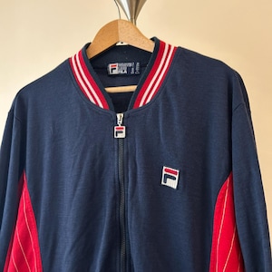 Puede incluir: Una chaqueta deportiva azul marino con cremallera y cuello acanalado a rayas rojas y blancas. La chaqueta incluye un parche con el logotipo de Fila en el pecho izquierdo, un tirador de cremallera con marca y paneles laterales rojos con rayas finas.