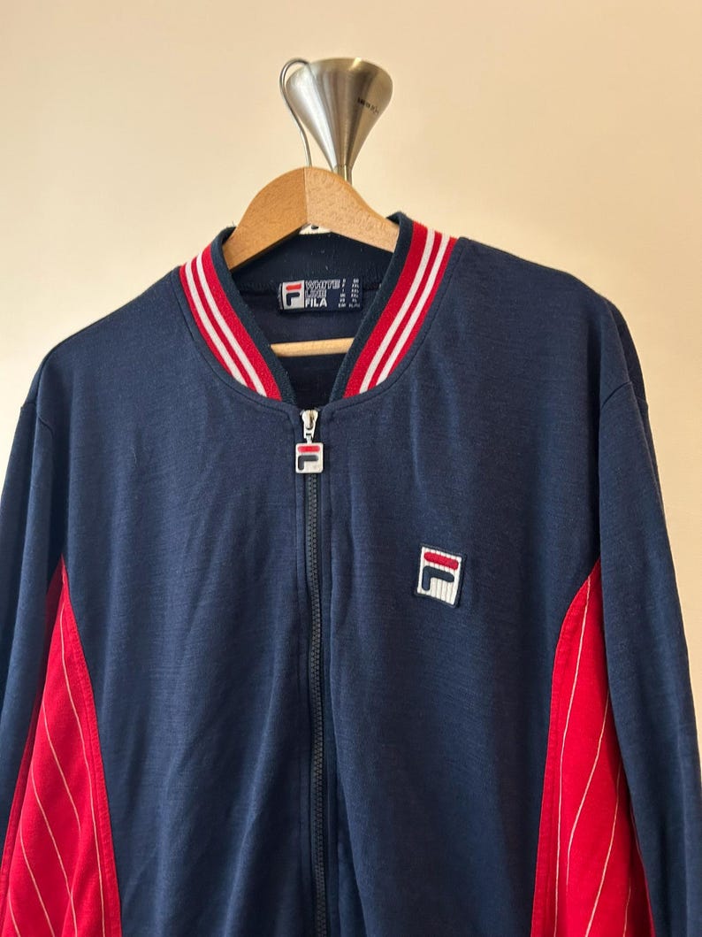 Puede incluir: Una chaqueta deportiva azul marino con cremallera y cuello acanalado a rayas rojas y blancas. La chaqueta incluye un parche con el logotipo de Fila en el pecho izquierdo, un tirador de cremallera con marca y paneles laterales rojos con rayas finas.