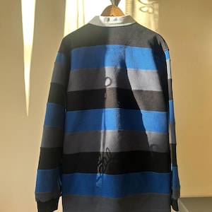 May include: A long-sleeved rugby shirt with horizontal stripes in black, blue, and gray. The shirt has a white collar and is displayed on a wooden hanger. The fabric appears to be a soft, fleece-like material.