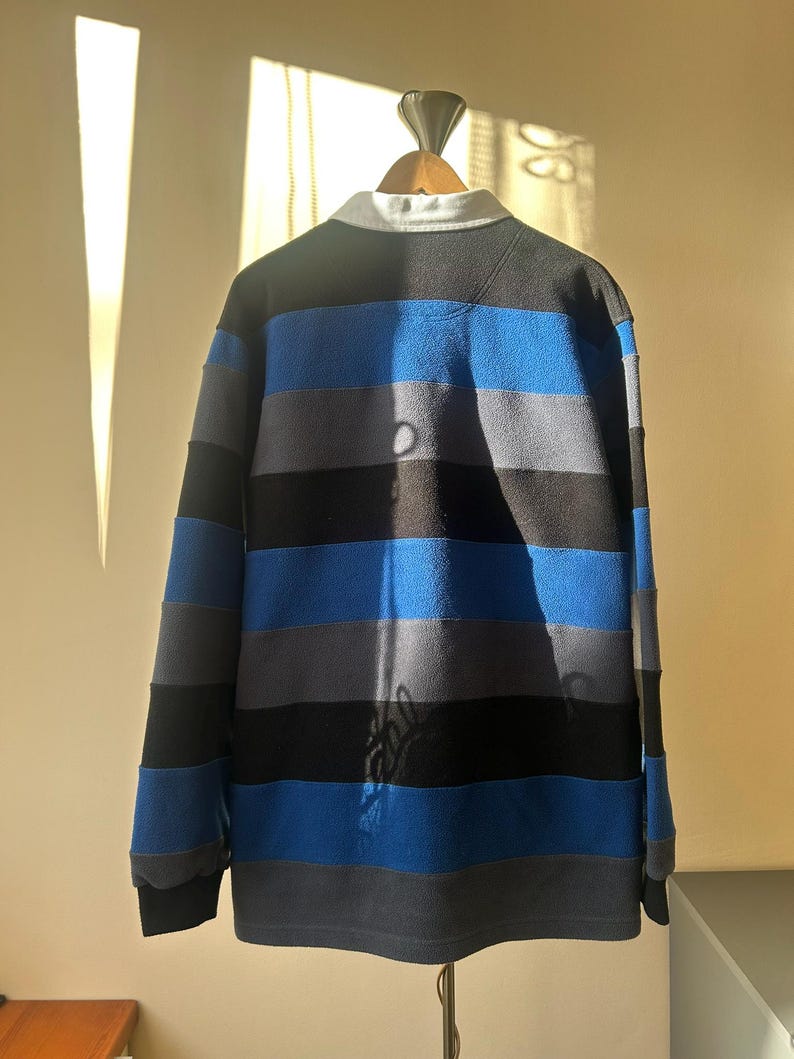 May include: A long-sleeved rugby shirt with horizontal stripes in black, blue, and gray. The shirt has a white collar and is displayed on a wooden hanger. The fabric appears to be a soft, fleece-like material.