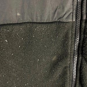 Pu&ograve; includere: Primo piano di una giacca in pile nera con cerniera a tutta lunghezza. La parte superiore &egrave; in tessuto nero liscio, mentre la sezione inferiore &egrave; in pile strutturato. La cerniera &egrave; argentata. La giacca sembra usurata.