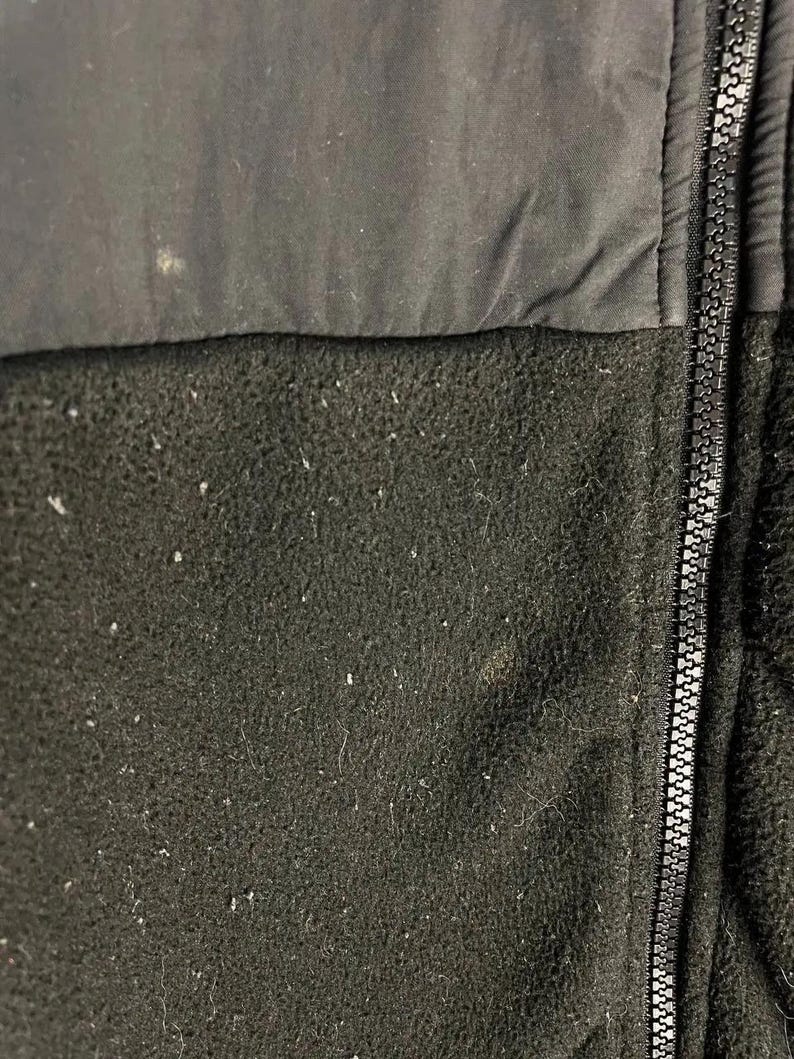 Pu&ograve; includere: Primo piano di una giacca in pile nera con cerniera a tutta lunghezza. La parte superiore &egrave; in tessuto nero liscio, mentre la sezione inferiore &egrave; in pile strutturato. La cerniera &egrave; argentata. La giacca sembra usurata.