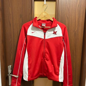 Peut inclure: Veste de survêtement zippée rouge et blanche avec le logo Nike et la lettre "M" sur la poitrine. La veste a une bande blanche le long des manches et sur la poitrine. La veste est suspendue à un cintre en bois.
