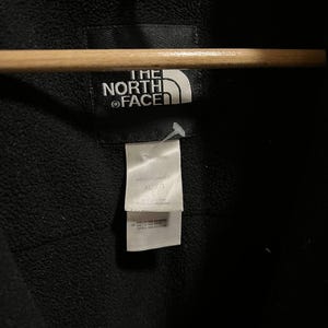 Pu&ograve; includere: Primo piano di una giacca nera con il logo bianco "THE NORTH FACE". Un'etichetta bianca recita "MENS/HOMMES XL/G". La giacca &egrave; appesa a un appendiabiti di legno.