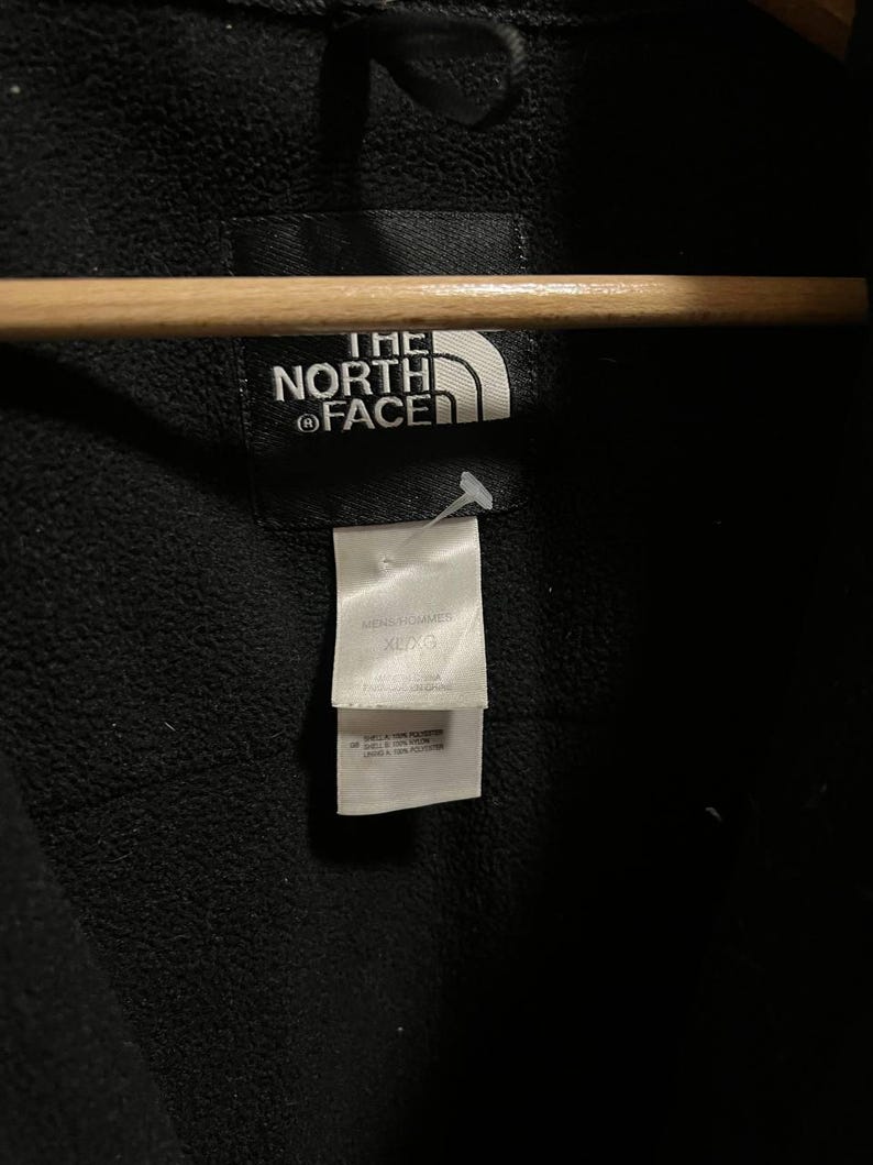 Pu&ograve; includere: Primo piano di una giacca nera con il logo bianco "THE NORTH FACE". Un'etichetta bianca recita "MENS/HOMMES XL/G". La giacca &egrave; appesa a un appendiabiti di legno.
