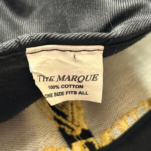 Pu&ograve; includere: Primo piano di un cappellino da baseball nero con un'etichetta che recita "THE MARQUE", "100% COTTON" e "ONE SIZE FITS ALL". Il cappellino ha un tessuto strutturato e ricami dorati.