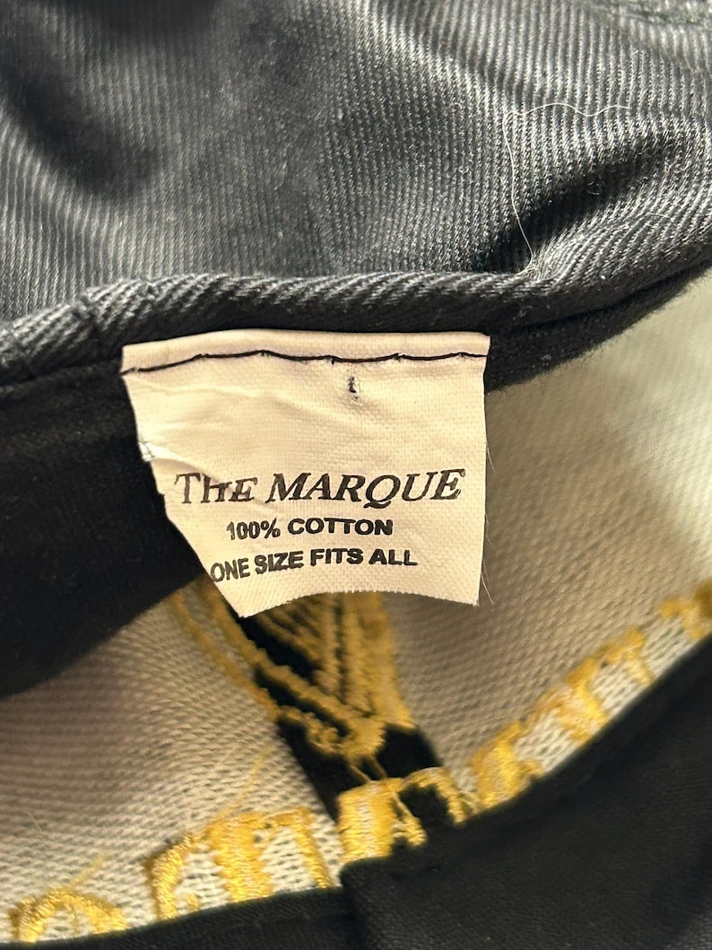Pu&ograve; includere: Primo piano di un cappellino da baseball nero con un'etichetta che recita "THE MARQUE", "100% COTTON" e "ONE SIZE FITS ALL". Il cappellino ha un tessuto strutturato e ricami dorati.