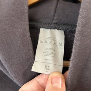 May include: Close-up of a dark gray hoodie with a tag showing washing instructions and the size XL. The tag is white with black text and symbols. The hoodie is hanging on a wooden hanger.