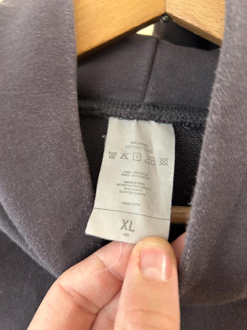 May include: Close-up of a dark gray hoodie with a tag showing washing instructions and the size XL. The tag is white with black text and symbols. The hoodie is hanging on a wooden hanger.