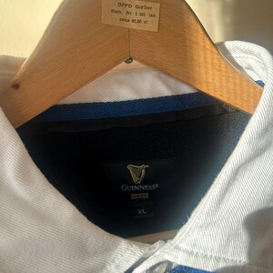 May include: A blue and white collared shirt with the Guinness logo. The shirt is on a wooden hanger. The shirt has a button-down collar and a blue body. The tag on the hanger has text.