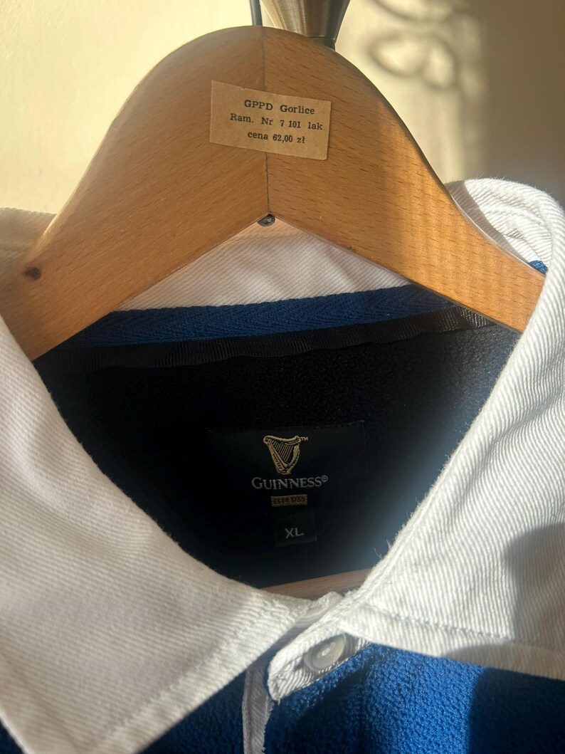May include: A blue and white collared shirt with the Guinness logo. The shirt is on a wooden hanger. The shirt has a button-down collar and a blue body. The tag on the hanger has text.