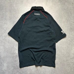 May include: Black short-sleeved polo shirt with a black collar and red trim. The word "Guinness" is embroidered in white on the back of the collar. The Guinness logo is on the sleeve.