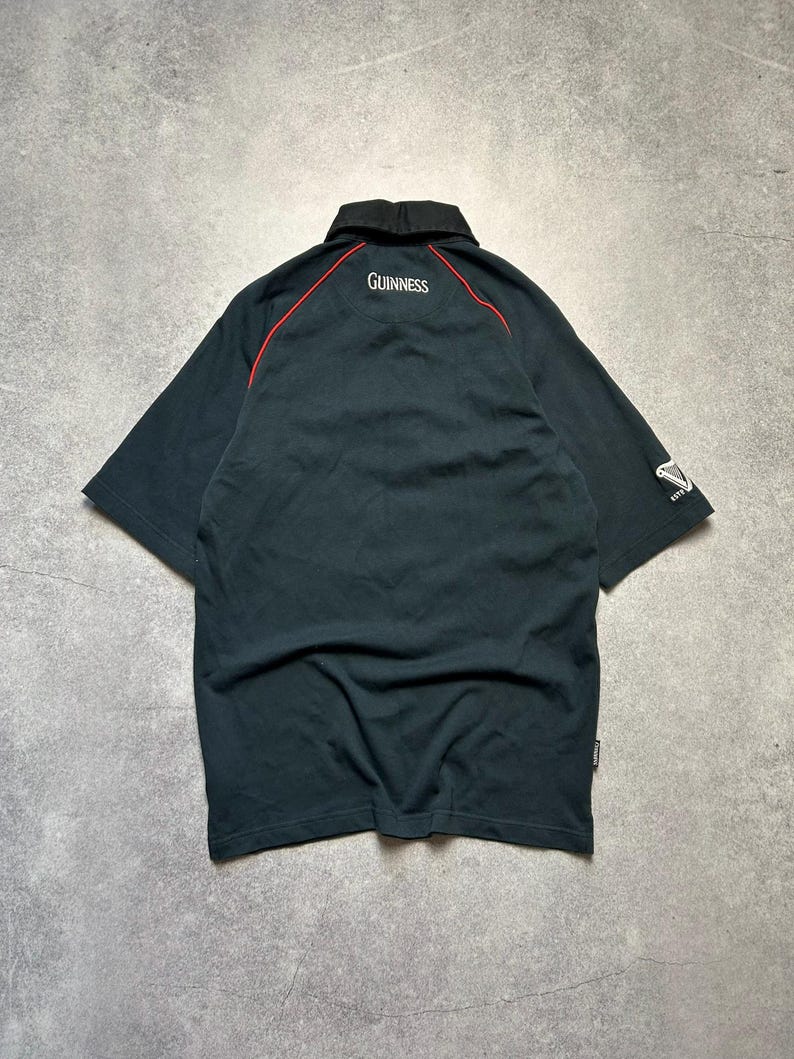 May include: Black short-sleeved polo shirt with a black collar and red trim. The word "Guinness" is embroidered in white on the back of the collar. The Guinness logo is on the sleeve.