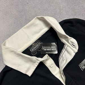 Puede incluir: Camiseta de rugby negra con cuello y tapeta de botones color crema. La camiseta presenta el logotipo de Guinness y una etiqueta que dice "Guinness + Rugby The Perfect Match Official Merchandise."