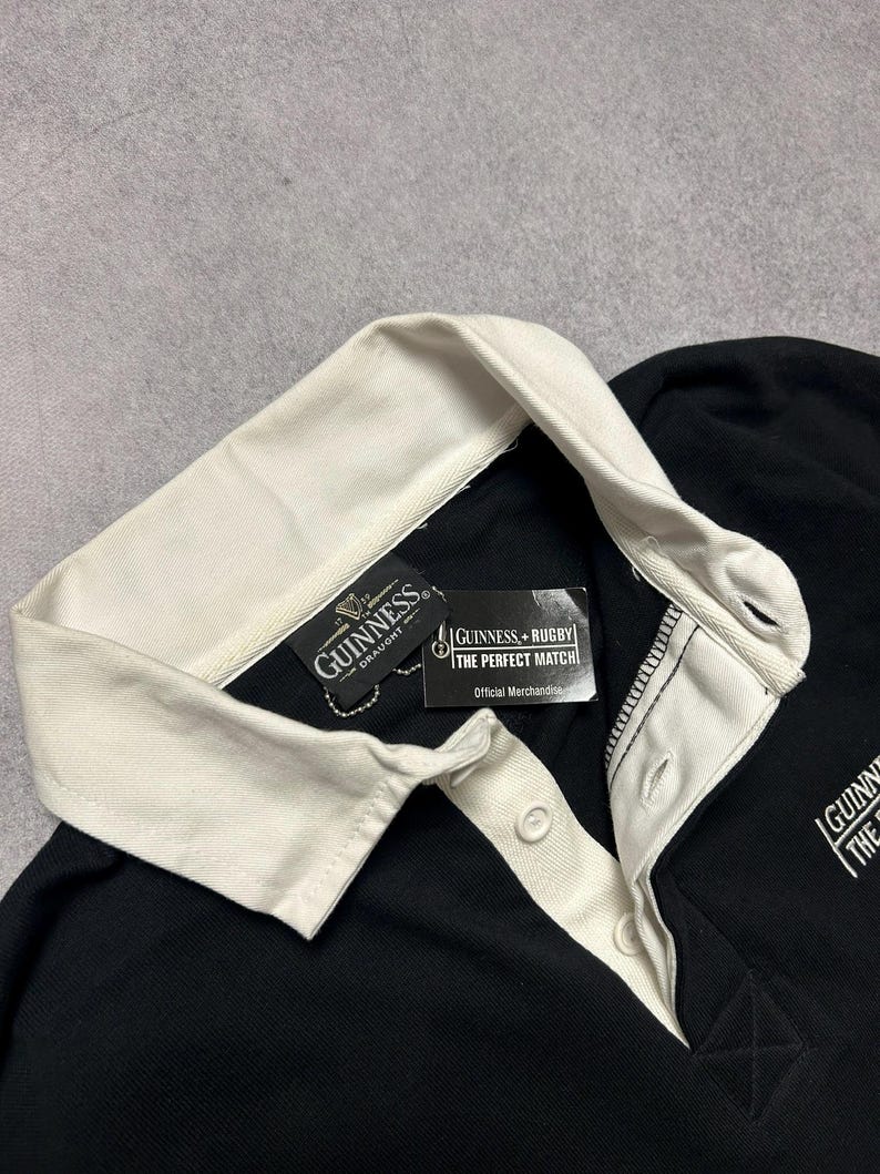 Puede incluir: Camiseta de rugby negra con cuello y tapeta de botones color crema. La camiseta presenta el logotipo de Guinness y una etiqueta que dice "Guinness + Rugby The Perfect Match Official Merchandise."
