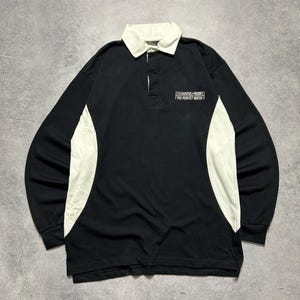 Puede incluir: Camiseta de rugby negra de manga larga con cuello y paneles laterales blancos. La camiseta presenta el texto "Guinness Rugby The Perfect Match" en el pecho. Tiene una tapeta abotonada.
