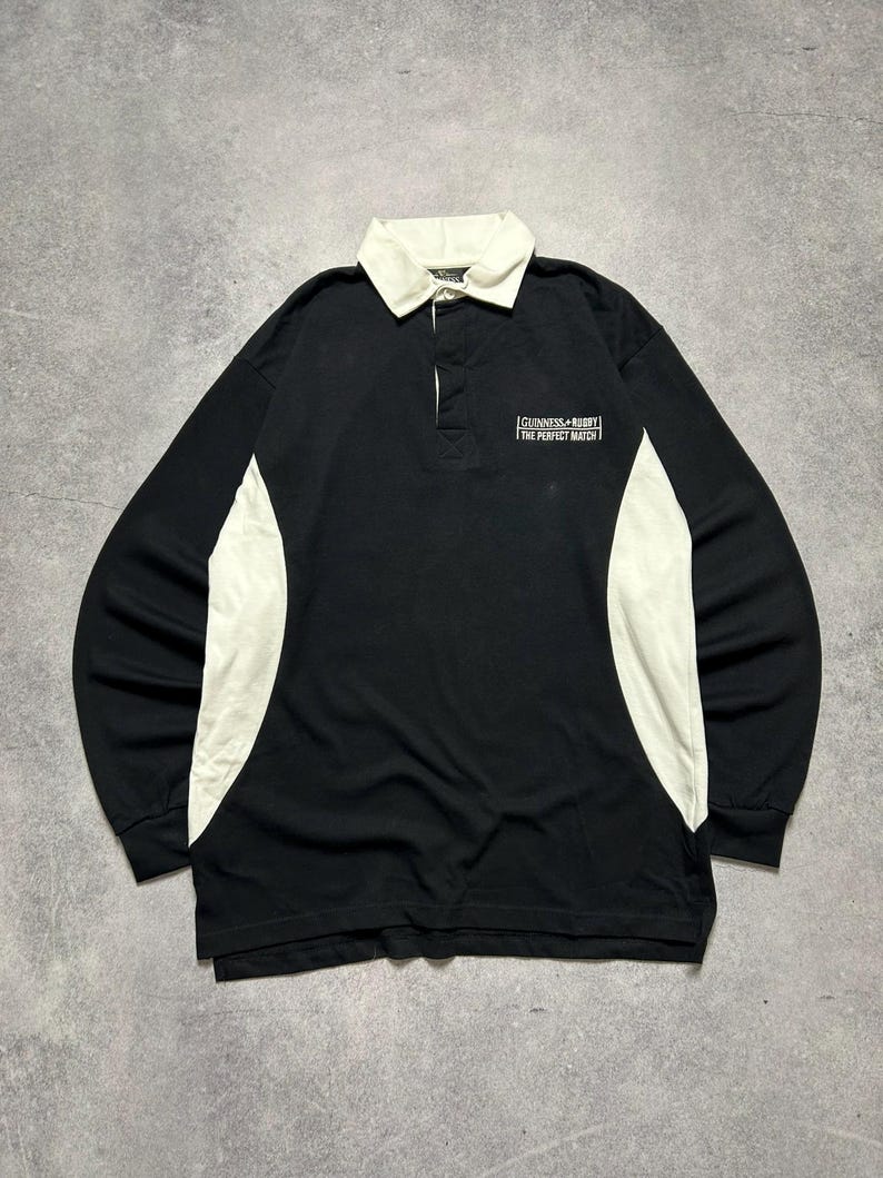 Puede incluir: Camiseta de rugby negra de manga larga con cuello y paneles laterales blancos. La camiseta presenta el texto "Guinness Rugby The Perfect Match" en el pecho. Tiene una tapeta abotonada.