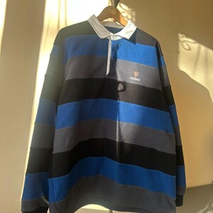 May include: A long-sleeved rugby shirt with horizontal stripes in blue, gray, and black. The shirt has a white collar and a small Guinness logo on the chest. The shirt is hanging on a wooden hanger.