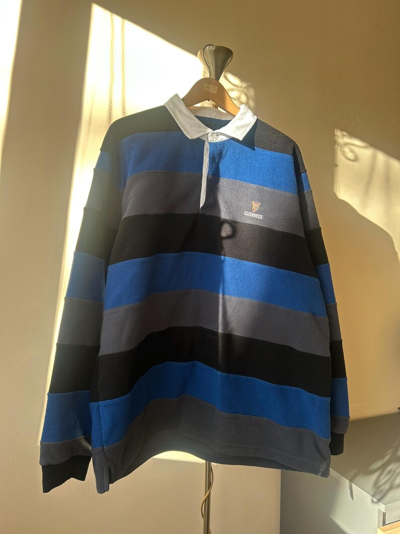 May include: A long-sleeved rugby shirt with horizontal stripes in blue, gray, and black. The shirt has a white collar and a small Guinness logo on the chest. The shirt is hanging on a wooden hanger.