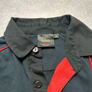 May include: A navy blue polo shirt with black collar and accents. The shirt features a red stripe detail and the "Official Guinness Merchandise" logo on the label. The shirt has two black buttons.