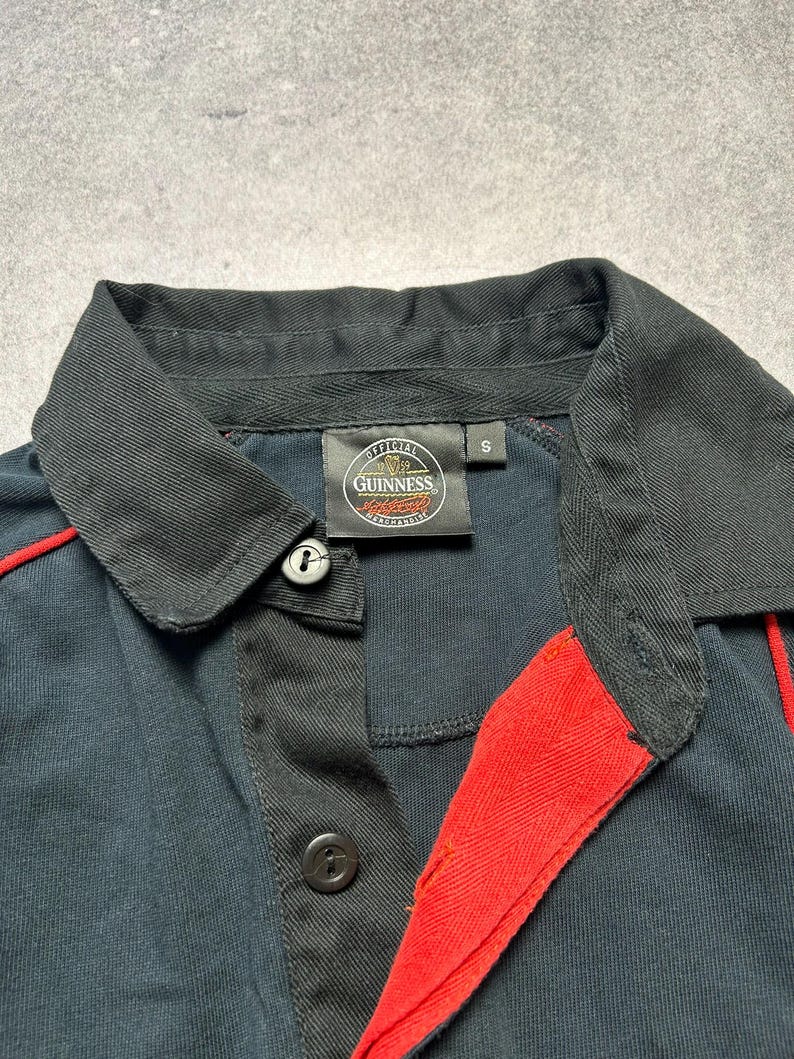 May include: A navy blue polo shirt with black collar and accents. The shirt features a red stripe detail and the "Official Guinness Merchandise" logo on the label. The shirt has two black buttons.