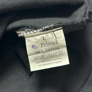 Puede incluir: Primer plano de una etiqueta de prenda negra con la marca "Pelmark" y el texto "100% COTTON". La etiqueta tambi&eacute;n incluye instrucciones de lavado y la talla "L".