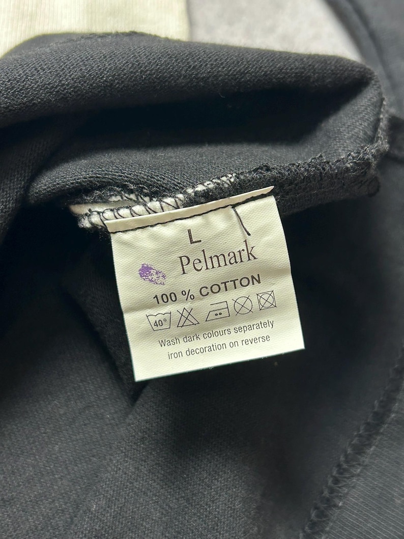 Puede incluir: Primer plano de una etiqueta de prenda negra con la marca "Pelmark" y el texto "100% COTTON". La etiqueta tambi&eacute;n incluye instrucciones de lavado y la talla "L".