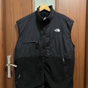 Peut inclure: Veste sans manches noire avec col montant et fermeture éclair intégrale. La partie supérieure est en matière lisse et matelassée, tandis que la moitié inférieure est en polaire douce. Le logo The North Face est sur la poitrine gauche. La veste est suspendue à un cintre en bois.