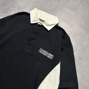 Puede incluir: Camiseta de rugby negra de manga larga con cuello blanco y panel lateral blanco. La camiseta presenta el texto "Guinness + Rugby The Perfect Match" en blanco en el pecho. La camiseta es de material suave.
