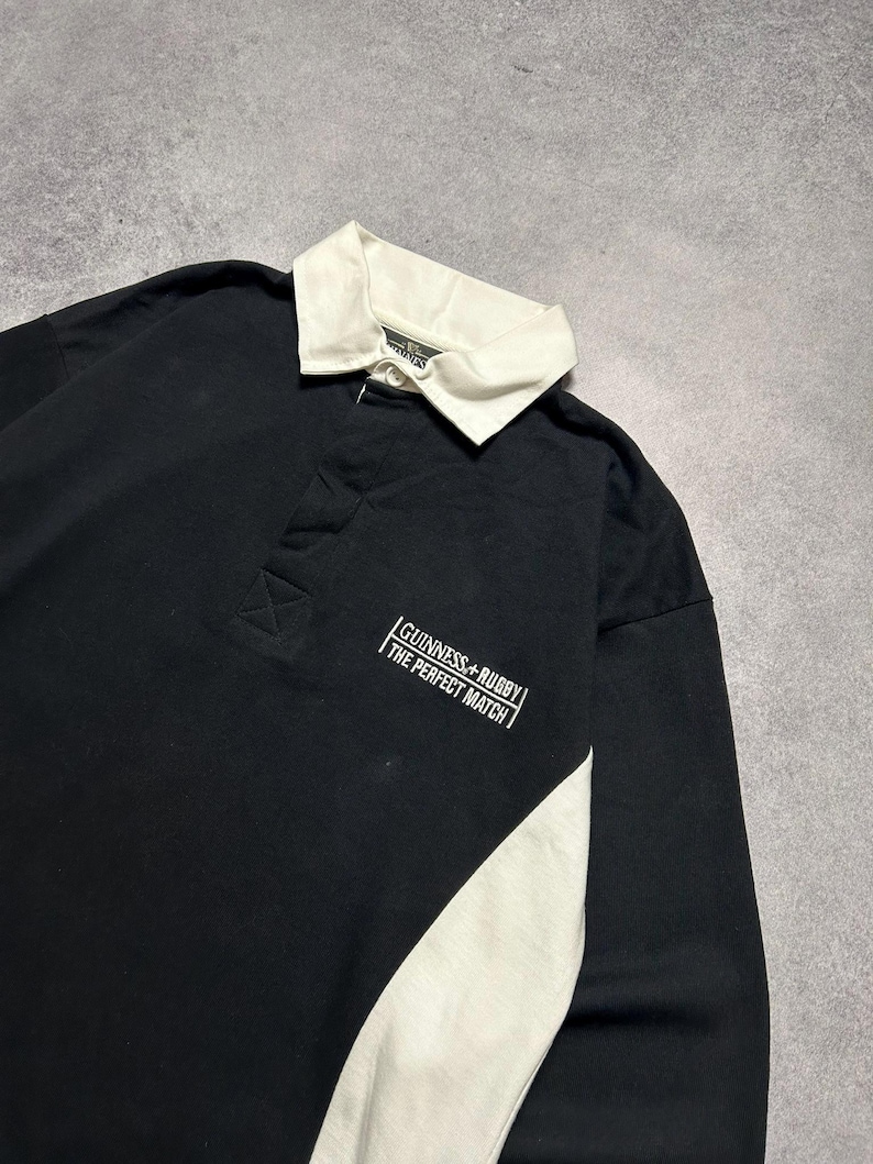 Puede incluir: Camiseta de rugby negra de manga larga con cuello blanco y panel lateral blanco. La camiseta presenta el texto "Guinness + Rugby The Perfect Match" en blanco en el pecho. La camiseta es de material suave.
