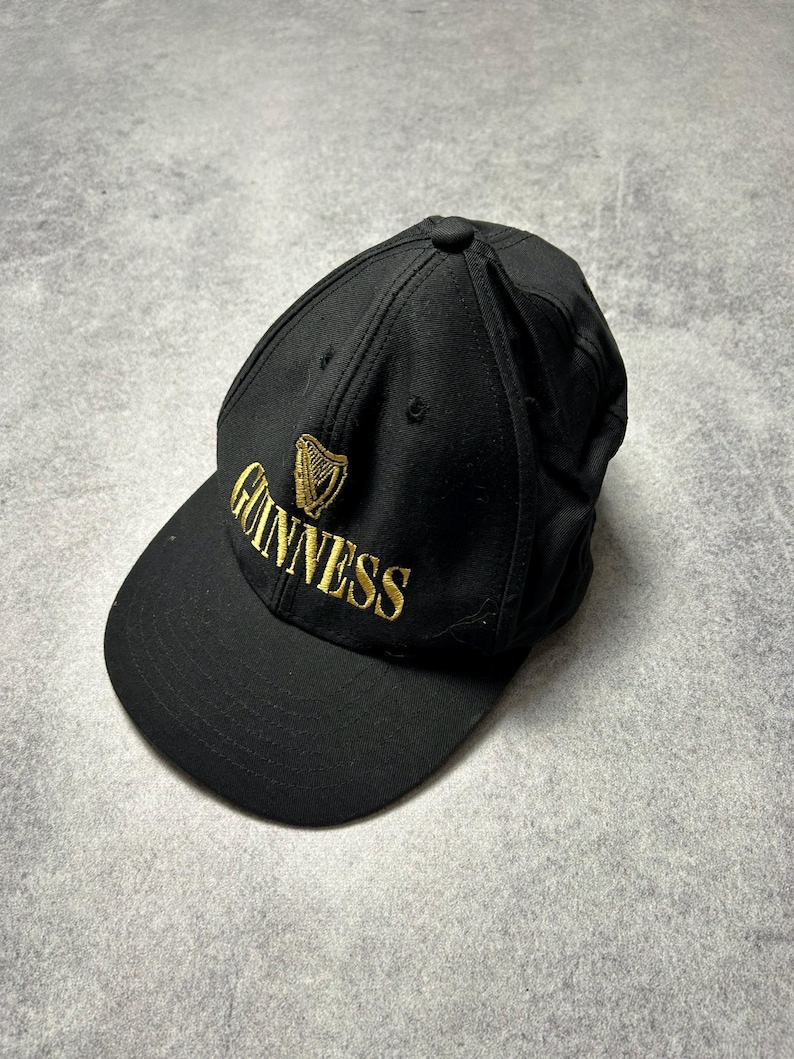 Pu&ograve; includere: Cappellino da baseball nero con il logo Guinness ricamato in oro. Il cappellino ha una visiera curva e un bottone sulla parte superiore. Il logo Guinness presenta un'arpa e il nome del marchio.