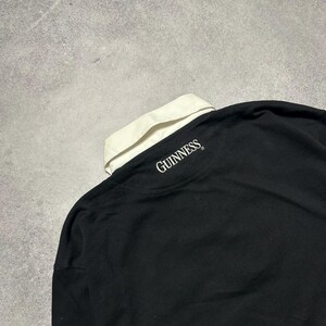 Puede incluir: Camisa negra de manga larga con cuello blanco y detalles blancos. La palabra "GUINNESS" est&aacute; impresa en blanco en la parte posterior del cuello. La camisa est&aacute; hecha de un material suave.