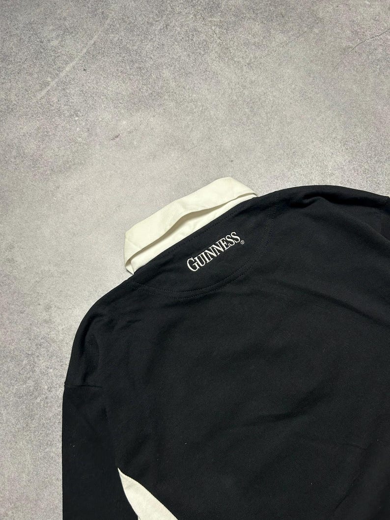 Puede incluir: Camisa negra de manga larga con cuello blanco y detalles blancos. La palabra "GUINNESS" est&aacute; impresa en blanco en la parte posterior del cuello. La camisa est&aacute; hecha de un material suave.