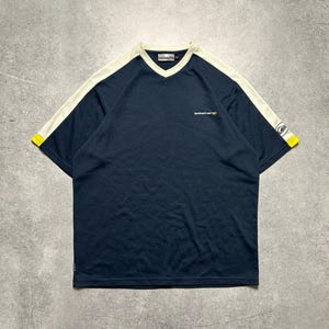 May include: Navy blue short-sleeve shirt with a V-neck and white stripes on the shoulders. The sleeves have yellow accents. The shirt has the Quiksilver logo on the chest and sleeve.