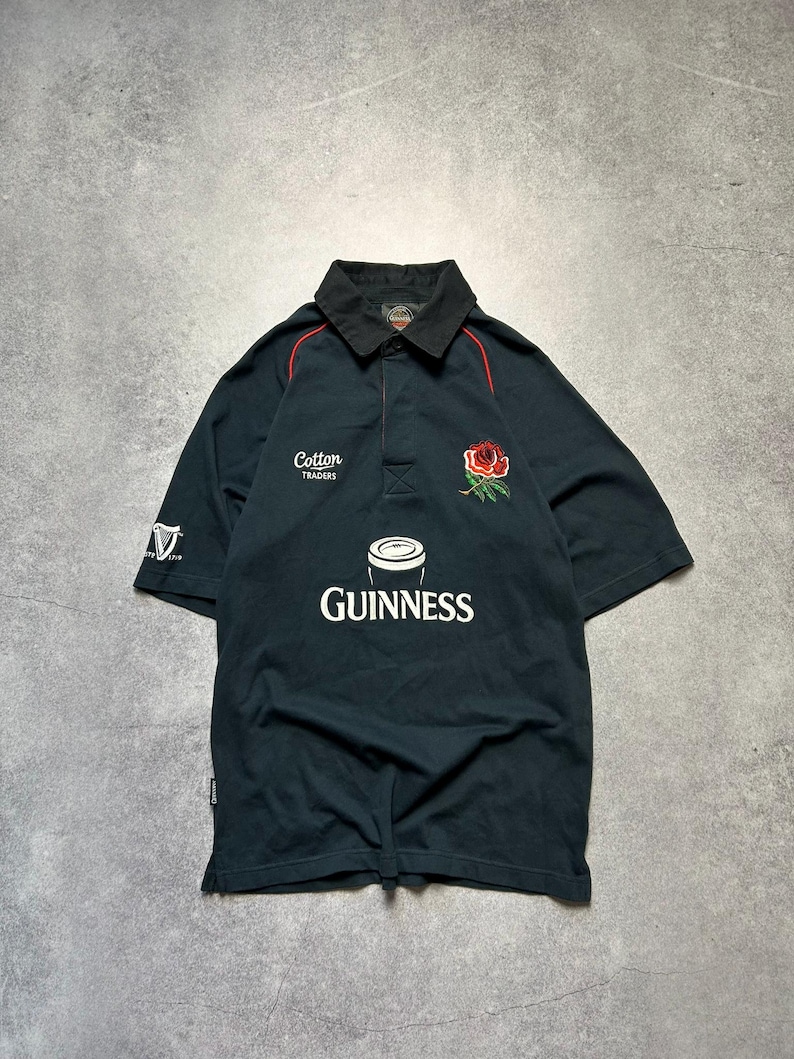 May include: Black rugby shirt with a black collar and red trim. The shirt features the Guinness logo, a red rose emblem, and the text "Cotton Traders". The left sleeve has a white "V" logo.