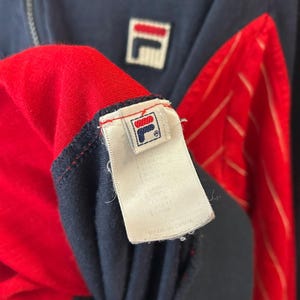 Puede incluir: Una chaqueta con cremallera azul marino y roja que presenta un parche con el logotipo de Fila en blanco en el pecho. La etiqueta interior muestra el logotipo de Fila y el texto Made in China. Incluye paneles rojos con finas rayas blancas y un tirador de cremallera de metal plateado.