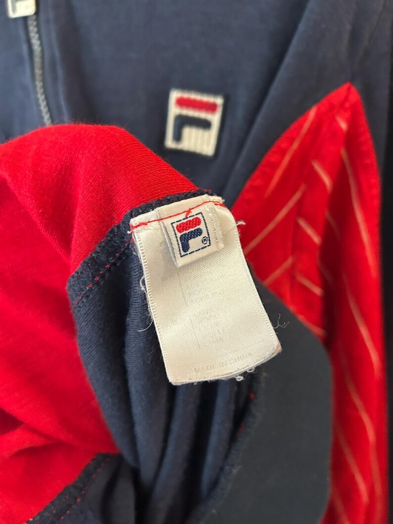 Puede incluir: Una chaqueta con cremallera azul marino y roja que presenta un parche con el logotipo de Fila en blanco en el pecho. La etiqueta interior muestra el logotipo de Fila y el texto Made in China. Incluye paneles rojos con finas rayas blancas y un tirador de cremallera de metal plateado.