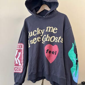 May include: Dark gray hoodie with the text "lucky me see Ghosts" in yellow, a pink heart with the word "feel", and flame graphics. The left sleeve has a red and white graphic. The hoodie has a hood and long sleeves.