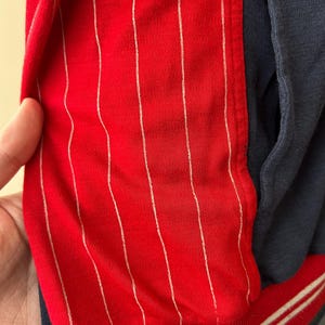 Puede incluir: Una chaqueta que presenta un panel rojo vibrante con finas rayas verticales blancas, contrastado por una secci&oacute;n de color azul marino oscuro s&oacute;lido. La prenda incluye un dobladillo acanalado en la parte inferior, azul marino con dos rayas blancas.