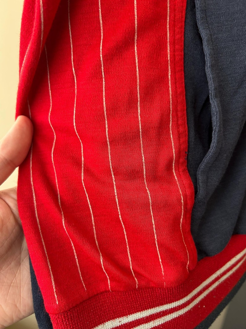 Puede incluir: Una chaqueta que presenta un panel rojo vibrante con finas rayas verticales blancas, contrastado por una secci&oacute;n de color azul marino oscuro s&oacute;lido. La prenda incluye un dobladillo acanalado en la parte inferior, azul marino con dos rayas blancas.